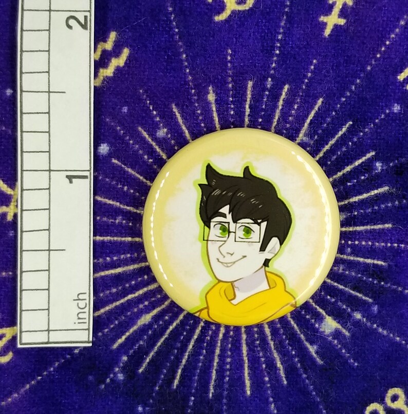 Puede incluir: Un bot&oacute;n amarillo con una ilustraci&oacute;n de dibujos animados de una persona con ojos verdes y cabello casta&ntilde;o que lleva una sudadera con capucha amarilla. La persona est&aacute; sonriendo.