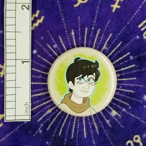 Puede incluir: Un bot&oacute;n de 2,5 cm de di&aacute;metro con una ilustraci&oacute;n de dibujos animados de una persona con cabello casta&ntilde;o, que lleva gafas y una bufanda marr&oacute;n. El fondo es de color amarillo verdoso con un halo azul claro alrededor de la persona.
