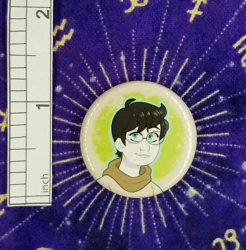 Puede incluir: Un bot&oacute;n de 2,5 cm de di&aacute;metro con una ilustraci&oacute;n de dibujos animados de una persona con cabello casta&ntilde;o, que lleva gafas y una bufanda marr&oacute;n. El fondo es de color amarillo verdoso con un halo azul claro alrededor de la persona.