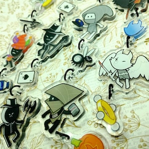 May include: A collection of colorful, cartoon-style character keychains. The characters include a pumpkin, a bee, a ghost, a knight, a mushroom, and a black cat. The keychains are made of clear acrylic and have a glossy finish.