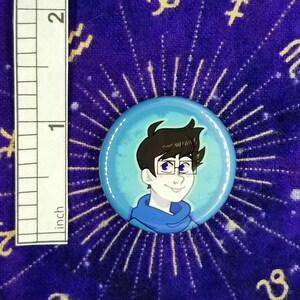 Puede incluir: Un bot&oacute;n azul y blanco con una ilustraci&oacute;n de dibujos animados de una persona con cabello casta&ntilde;o que lleva gafas y un su&eacute;ter azul.