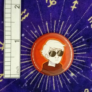 Puede incluir: Un bot&oacute;n rojo con una ilustraci&oacute;n de dibujos animados de una persona con cabello blanco que lleva gafas de sol. La persona lleva un su&eacute;ter marr&oacute;n y rojo.