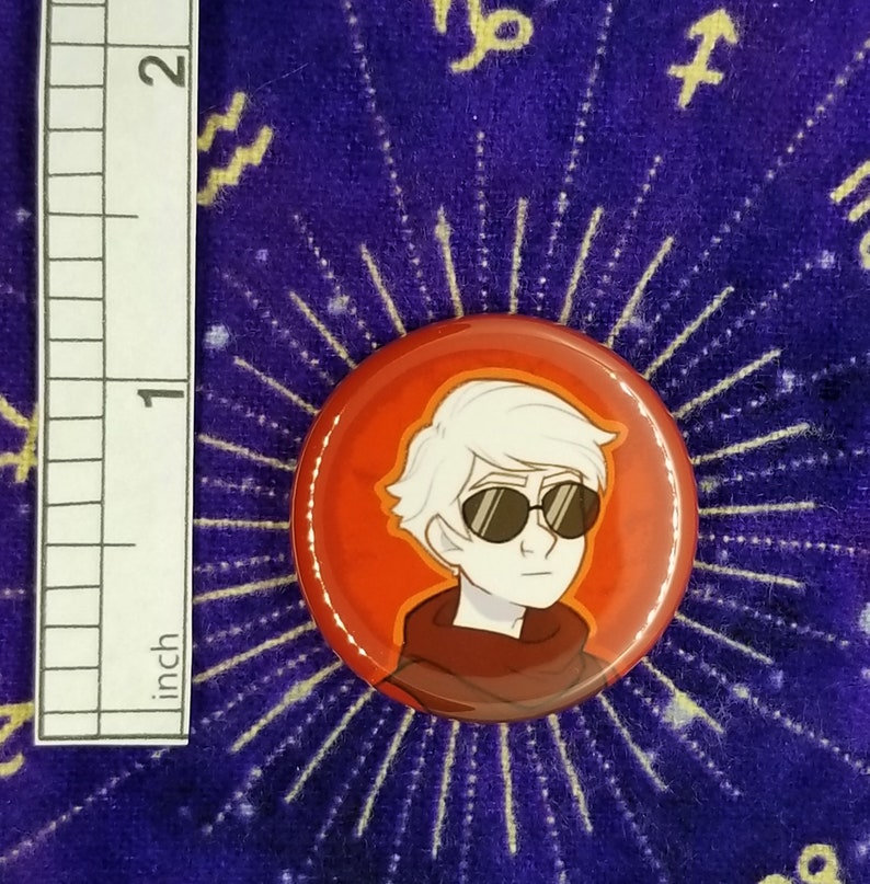 Puede incluir: Un bot&oacute;n rojo con una ilustraci&oacute;n de dibujos animados de una persona con cabello blanco que lleva gafas de sol. La persona lleva un su&eacute;ter marr&oacute;n y rojo.