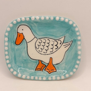 Può includere: Un piatto quadrato in ceramica con un disegno dipinto a mano di un'oca bianca con becco e zampe arancioni. L'oca è posta su uno sfondo azzurro. Il bordo del piatto è bianco con piccoli punti distanziati.