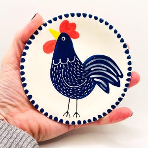 Puede incluir: Plato pequeño y redondo de cerámica con un diseño de gallo pintado a mano. El gallo es azul marino con una cresta roja y un pico amarillo. El plato tiene un fondo blanco y un borde azul punteado. El plato se sostiene en una mano.