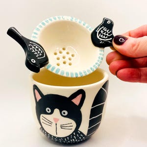 Könnte beinhalten: Ein Keramik-Teebecher und Infuser-Set. Der Becher hat ein schwarzes Katzengesicht-Design. Der Infuser ist weiß mit hellblauer Umrandung und zwei schwarzen Vogelgriffen. Das Set ist zum Aufbrühen von losem Tee konzipiert.