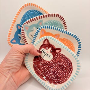 Peut inclure: Un ensemble de quatre petits plats en céramique, chacun orné d'une illustration de chat peinte à la main. Les plats sont colorés dans des tons bleu foncé, bleu clair, orange et rouge profond, avec des bordures à motifs assorties.