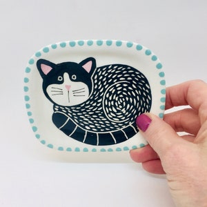 Op de afbeelding: Een witte keramische schaal met een blauwe gestippelde rand heeft een zwart-witte kat die in slaappositie is opgerold. De kat heeft een roze neus en witte snorharen.
