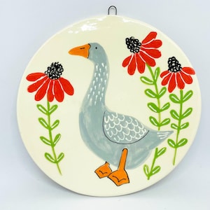 May include: Round ceramic wall hanging featuring a hand-painted goose with gray feathers, orange beak and feet, and red flowers with black centers. The background is off-white, and the piece has a metal hook for hanging.