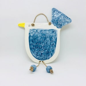 Puede incluir: Un pájaro de cerámica blanca con un cuerpo estampado en azul y blanco y un pico amarillo. El pájaro tiene una cuerda que cuelga de su cuerpo con cuentas azules en el extremo.