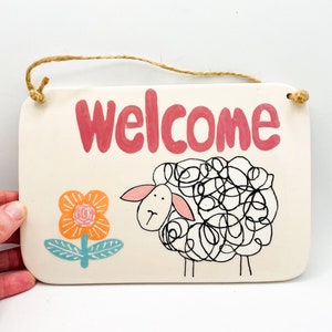 May include: Cream-coloured rectangular sign with the word "Welcome" in red lettering. A whimsical sheep illustration and a flower are on the sign. A jute rope is attached for hanging.