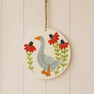 May include: A circular white wall hanging featuring a hand-painted gray goose with orange feet and beak, flanked by two tall red flowers with green stems and leaves. The piece is suspended by a thin brown twine loop attached to the top edge.