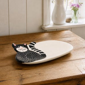 Può includere: Un vassoio in ceramica a forma di gatto, caratterizzato da una figura felina con motivo bianco e nero su un lato e una superficie liscia color crema sull'altro. Il design del gatto include orecchie nere, un naso rosa e una coda a strisce.