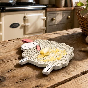 May include: A white ceramic grater shaped like a sheep with a textured surface for grating. The sheep has a smooth head with pink ears, black outlines for facial features, and four short legs. A small piece of ginger is being grated on the surface.
