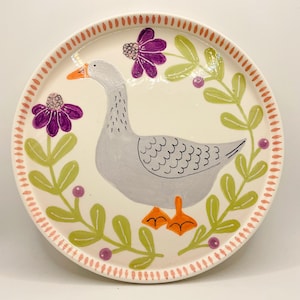 May include: A decorative ceramic plate featuring a hand-painted goose design. The goose is gray with an orange beak and feet, surrounded by purple flowers and green leaves. The plate's rim has a dotted pink border.