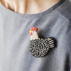 Może przedstawiać: Ceramiczna broszka w kształcie kury z białą głową, czerwonym grzebieniem i dzwonkami oraz czarnym tułowiem z białymi cętkami. Kura jest przedstawiona z profilu, zwrócona w lewo, z żółtym dziobem i gładkim, błyszczącym wykończeniem powierzchni.
