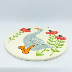 May include: A round, off-white ceramic plaque featuring a hand-painted illustration of a grey goose with orange feet. The goose is positioned between two vertical stems with green leaves and bright red, daisy-like flowers on a light background.