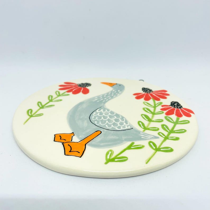 May include: A round, off-white ceramic plaque featuring a hand-painted illustration of a grey goose with orange feet. The goose is positioned between two vertical stems with green leaves and bright red, daisy-like flowers on a light background.