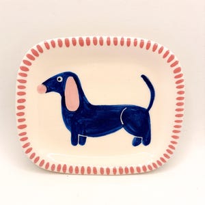 Könnte beinhalten: Rechteckige Keramikschale mit cremefarbenem Grund und handgemalter Illustration eines blauen Dackels. Die Schale hat einen rosa gepunkteten Rand. Der Hund hat rosa Ohren und eine rosa Nase.