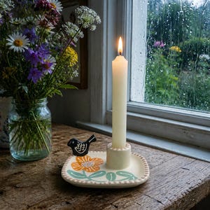 May include: A cream-colored ceramic candle holder with a square base featuring a painted orange flower, green leaves, and a small black bird figure. A single lit white taper candle stands in the center holder, which has a dotted rim design.