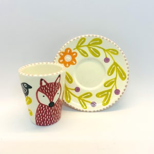 Könnte beinhalten: Ein weißes Keramik-Teetassen- und Untertassen-Set mit handbemaltem Design eines Fuchses, eines Vogels und floraler Elemente. Tasse und Untertasse haben einen rosa Rand.