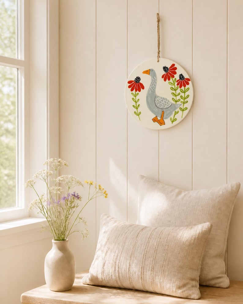 May include: A circular white wall hanging featuring a hand-painted gray goose with orange feet and beak, flanked by two tall red flowers with green stems and leaves. The piece is suspended by a thin brown twine loop attached to the top edge.