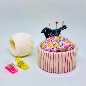 May include: A ceramic pin cushion with a floral fabric top and a striped base. A black bird-shaped pin holder and a ball of white thread are nearby. Two sewing clips in pink and yellow are also present.