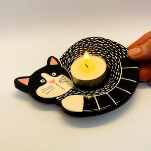 Może przedstawiać: Ceramiczny świecznik w kształcie czarno-białego kota z małą białą świeczką typu tealight w środku. Kot jest zwinięty z ogonem wokół świeczki.