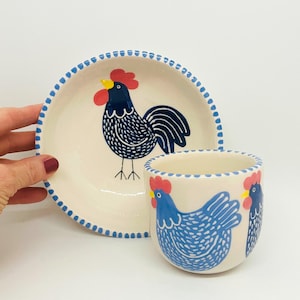 Puede incluir: Un juego de dos platos de cerámica con diseño de gallo y gallina. El plato más grande es un tazón con un gallo azul y blanco con cresta roja y pico amarillo. El plato más pequeño es una taza con dos gallinas azules y blancas con crestas rojas y picos amarillos.