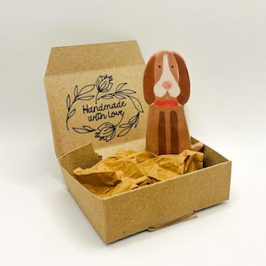Puede incluir: Una figurita de perro de cerámica marrón y blanca en una caja de cartón marrón con una etiqueta blanca que dice "Handmade with love".