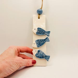 Pode incluir: Um pendurador de parede de cerâmica com três peixes azuis e brancos presos a uma base retangular esbranquiçada. Uma conta azul e uma corda de juta no topo. Os peixes têm escamas e barbatanas detalhadas.