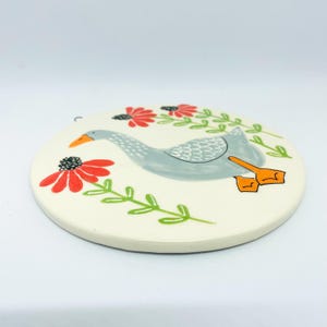 May include: A round, off-white ceramic coaster featuring a hand-painted illustration of a grey goose with an orange beak and feet. The goose is surrounded by stylized red flowers with black centers and green stems on a cream-colored background.