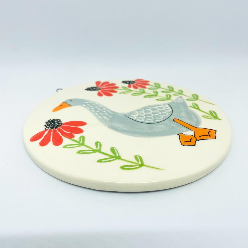 May include: A round, off-white ceramic coaster featuring a hand-painted illustration of a grey goose with an orange beak and feet. The goose is surrounded by stylized red flowers with black centers and green stems on a cream-colored background.