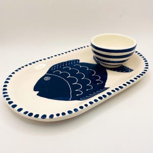 Puede incluir: Un juego de servir de cerámica con una fuente ovalada crema y un cuenco pequeño. La fuente tiene un diseño de pez azul oscuro y un borde punteado. El cuenco es blanco con rayas azules. Una adición encantadora a cualquier cocina.