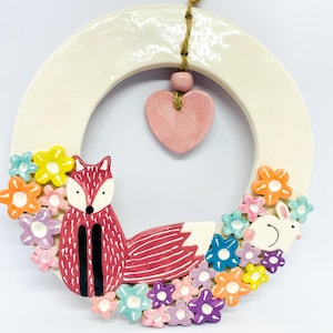 Peut inclure: Une couronne en céramique avec un renard rouge et un lapin blanc entourés de fleurs colorées. La couronne a un anneau crème avec un pendentif en forme de cœur rose et une ficelle de jute pour suspendre. Une pièce décorative.