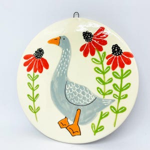 May include: A round ceramic wall plaque with a cream-colored background featuring a hand-painted gray goose with an orange beak and feet. The goose is surrounded by three stylized red flowers with black centers and green stems and leaves.