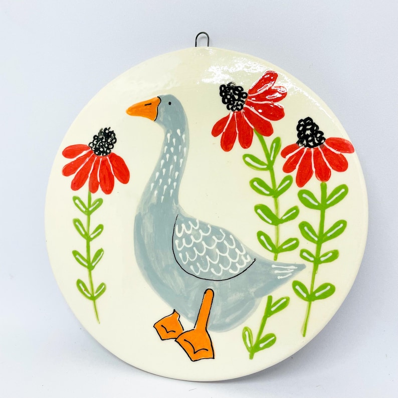 May include: A round ceramic wall plaque with a cream-colored background featuring a hand-painted gray goose with an orange beak and feet. The goose is surrounded by three stylized red flowers with black centers and green stems and leaves.
