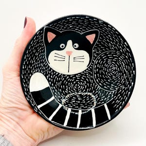 Może przedstawiać: Czarno-biały ceramiczny talerz z motywem kota. Kot ma czarną głowę z różowymi uszami i różowym nosem. Talerz ma teksturowane tło i pasiasty ogon. Unikalny element dekoracyjny.