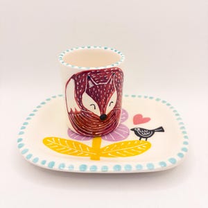 Könnte beinhalten: Ein Keramik-Tassen- und Untertassen-Set mit einem skurrilen Fuchsmotiv. Die Tasse zeigt eine Illustration eines roten Fuchses, und die Untertasse hat ein Blumen-, Vogel- und Herzmotiv. Das Set ist handbemalt mit blauen, gelben, rosa und schwarzen Akzenten.