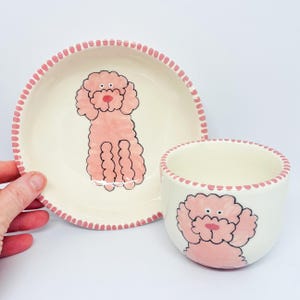 May include: A pink ceramic bowl and cup set with a pink poodle design. The bowl and cup have a pink and white dotted trim.