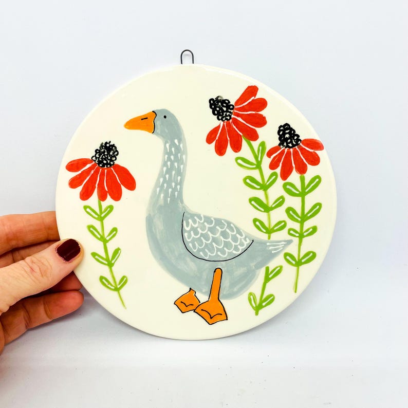 May include: A round white ceramic wall plaque featuring a hand-painted illustration of a grey goose with an orange beak and feet. The goose is surrounded by three stylized red flowers with black centers and green stems on a cream-colored background.