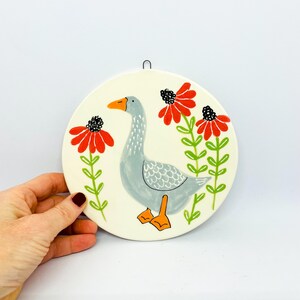 May include: A round ceramic wall plaque featuring a hand-painted illustration of a grey goose with an orange beak and feet. The goose is positioned between two tall green stems topped with vibrant red flowers with black centers.