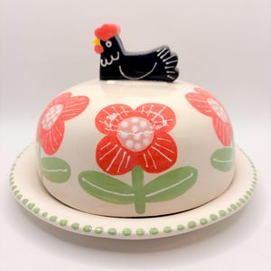 May include: A ceramic butter dish with a lid featuring a black chicken handle. The dish is decorated with red flowers and green leaves on a cream background. The base has a dotted green border.
