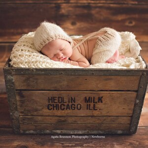 May include: A newborn baby wearing a knitted hat and trousers sleeps in a wooden crate with a white blanket. The crate has the text "Hedlin Milk Chicago Ill." printed on the front.