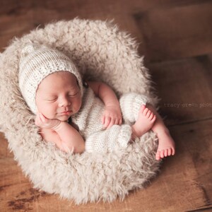 May include: A newborn baby wearing a white knitted hat and matching outfit is sleeping peacefully in a fluffy white  basket. The baby's face is visible and the baby's hand is resting on their chin.