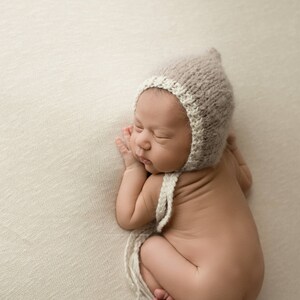 May include: A newborn baby wearing a beige knitted hat with a white trim, sleeping on a white textured blanket.