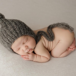 May include: A newborn baby wearing a gray knitted hat with a pom-pom on top and a matching scarf. The baby is sleeping on a white blanket.