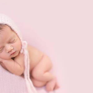 May include: A newborn baby girl sleeping peacefully on a pink blanket, wearing a pink knitted hat. The baby is curled up in a foetal position with her arms tucked under her chin.
