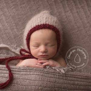 May include: A newborn baby wearing a white and burgundy knitted hat with a red bow. The baby is sleeping with their eyes closed and hands clasped together. The baby is lying on a grey textured blanket.