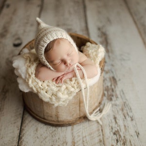 May include: A newborn baby wearing a cream knitted hat with a pointed tip, sleeping in a wooden bucket lined with a white crocheted blanket.